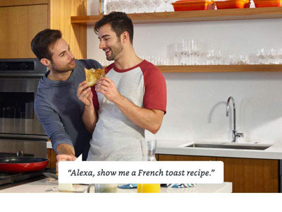   Two males in the kitchen, smiling at each.  A dialog box at the lower part of the picture contains texts reading as “Alexa, show me a French toast recipe.” 